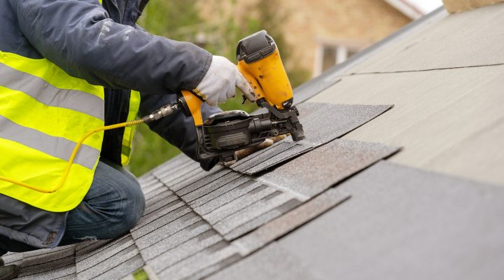 Unrecognizable roofer worker in special protective work wear and gloves, using air or pneumatic nail gun and installing asphalt or bitumen shingle on top of the new roof under construction residential building
